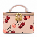 A structured top-handle handbag in light pink pebbled leather with a cherry print, featuring gold-tone hardware and a white protective wrap on the handle, shown against a plain white background.